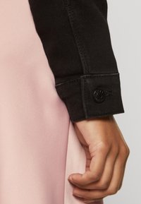 Hand resting beside pink fabric, wearing a black long-sleeve cuff with a black button.