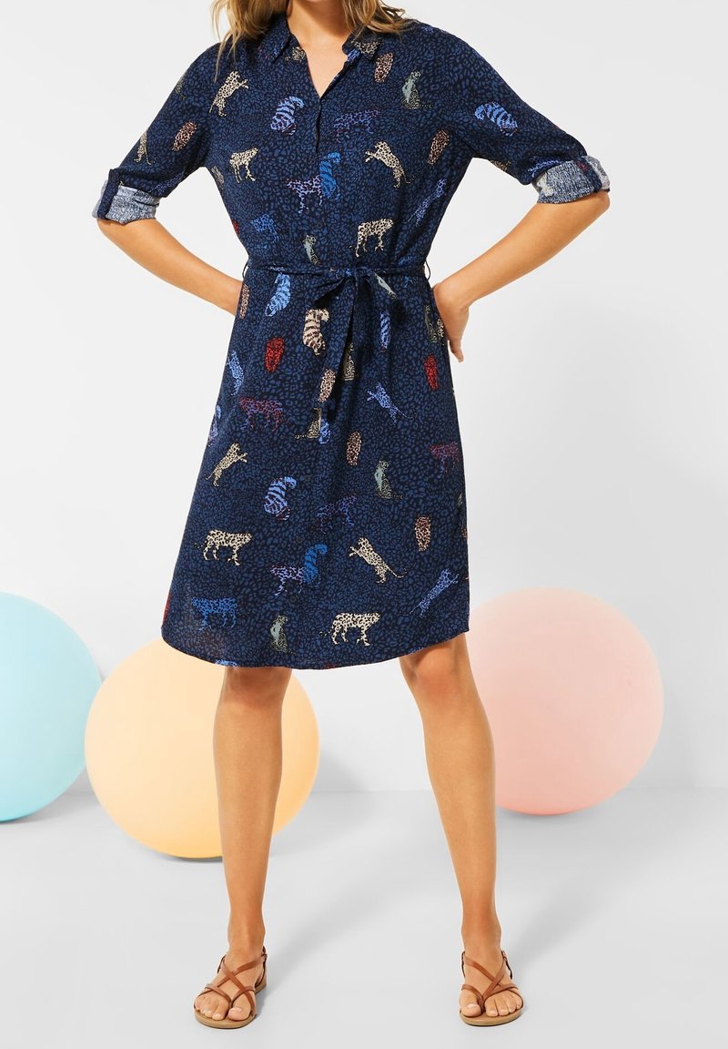 Femme portant une robe chemise bleu marine mi-longue avec des imprimés animaux colorés, manches retroussées, taille nouée et sandales marron à lanières, sur un fond uni.