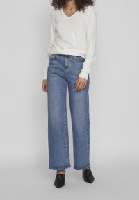 Jeans de pierna ancha azul con cintura alta, que presentan una textura de mezclilla y costuras sutiles. Combinados con un suéter blanco de escote en V y tacones negros.