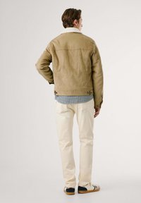 Bruine corduroy jas met een schapenvachtkraag, lange mouwen, met knopen bij de manchetten, gecombineerd met crèmekleurige broeken en sneakers. Achteraanzicht weergegeven.