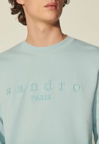 Světle modrá mikina s kulatým výstřihem a vyšitým nápisem "sandro PARIS" kontrastní nití. Hladká tkanina s uvolněným střihem.