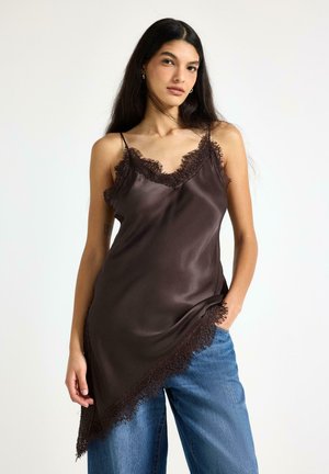 Donna con lunghi capelli scuri indossa un vestito sottoveste in satin marrone scuro con rifiniture in pizzo sopra jeans blu, in piedi con una mano in tasca.