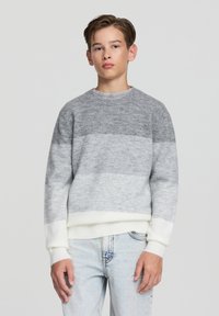 Szaro-biały sweter w paski z okrągłym dekoltem i ściągaczami, wykonany z miękkiego materiału, zestawiony z jasnoniebieskimi dżinsami.