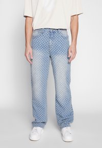 Man i ljusblå jeans med små vita kaktusmönster, kombinerat med vita sneakers och en löst sittande off-white t-shirt med kort ärm.