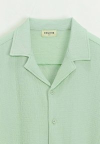 Chemise boutonnée vert clair en tissu texturé, avec un col et deux boutons à l'avant. L'étiquette indique "FRILIVIN, taille S."