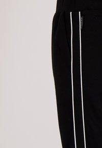 Zwarte broek van een textuurstof, met witte zijstrepen en een klein merkembleem op de tailleband. Close-up van het onderste gedeelte.