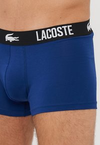 Boxers de algodón azul con una cinturilla elástica negra que presenta "LACOSTE" y un logo de cocodrilo blanco. Diseño ajustado y textura suave.