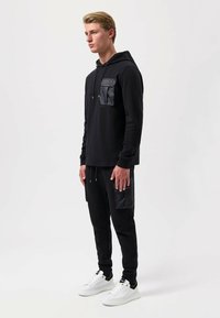 Felpa nera con tasca a toppa, abbinata a pantaloni neri a sigaretta con tasca laterale. Sneakers bianche completano l'outfit.