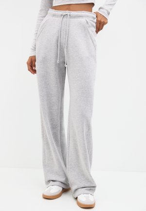 Femme portant un pantalon de jogging large gris clair avec taille à cordon et des baskets blanches, debout devant un fond blanc uni.