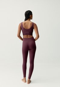 Mujer con el cabello trenzado que está descalza, vistiendo un sujetador deportivo de canalé burdeos a juego con leggings de tiro alto, frente a un fondo sencillo.
