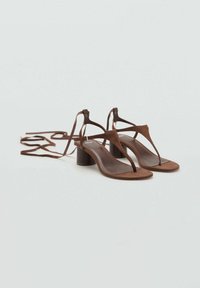 Brune ruskinds hæle sandaler med T-stroppedesign, kort blokhæl og lange snorebånd ved anklen. Glat fodseng med en minimalistisk stil.