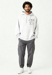 Felpa grigia con cappuccio, tasca a marsupio, grafica di un uccello nero e testo. Abbinata a pantaloni cargo grigi con tasche laterali e polsini elastici. Scarpe da ginnastica bianche.