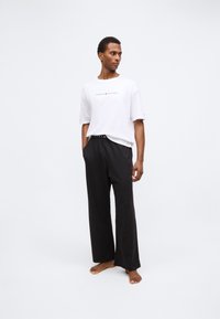 T-shirt blanc à manches courtes avec le logo "TOMMY HILFIGER", associé à un pantalon noir ample avec une taille logo, sur fond uni.