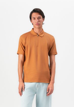 Ung mand iført en struktureret burnt orange polo med kort ærme og lynlås samt lysblå jeans, stående foran en ensfarvet hvid baggrund.