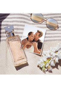 Eternity for Women doft i en klar glasflaska med ett silverlock, tillsammans med solglasögon, ett foto och vita blommor på ett mönstrat tyg.