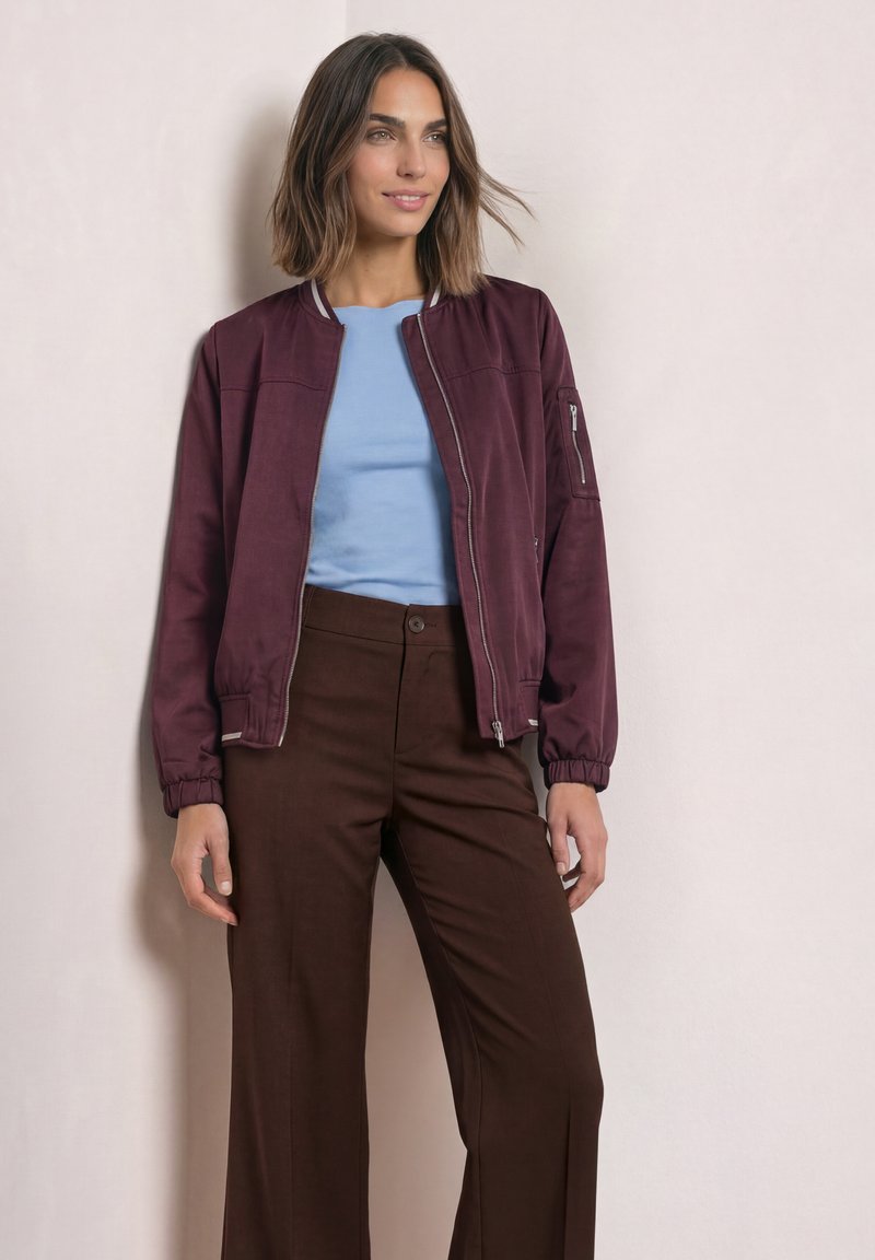 Woman with shoulder-length hair wearing a maroon bomber jacket, light blue shirt, and brown pants standing against beige wall.
