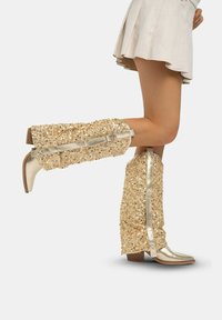 Botas de rodilla altas decoradas con lentejuelas doradas y un acabado metálico, que presentan un cierre lateral, un upper texturizado y un tacón apilado.