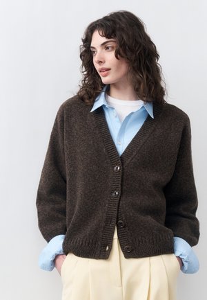 Vrouw met krullend donker haar, gekleed in een donkerbruine gebuttonde cardigan over een blauw overhemd met kraag en crèmegekleurde broek, handen in de zakken.