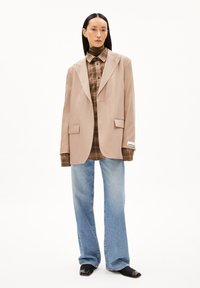 Blazer beige con rever a punta, indossato sopra una camicia a quadri marrone. Jeans svasati azzurri e scarpe nere a punta completano l'outfit.