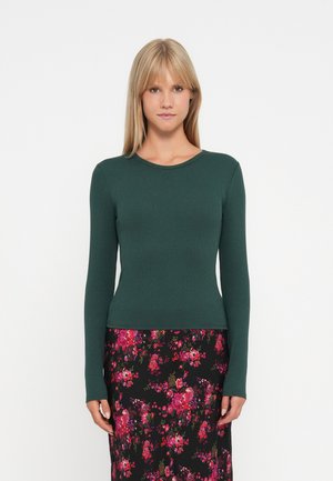 Maglietta verde a maniche lunghe con scollo rotondo, abbinata a una gonna nera floreale con motivi rosa, rossi e verdi.
