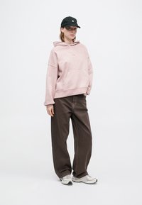 Prevelik roza hoodie s sprednjim žepom, v kombinaciji s širokimi rjavimi hlačami in sivimi športnimi copati. Črna kapa dopolnjuje sproščen videz.