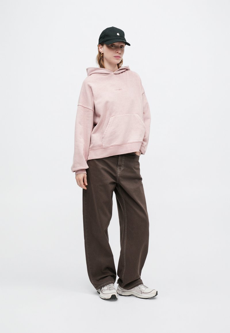 Übergrößte rosa Kapuzenjacke mit einer vorderen Tasche, kombiniert mit braunen weiten Hosen und grauen Turnschuhen. Eine schwarze Mütze rundet den lässigen Look ab.