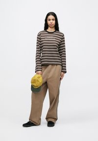 Fiatal nő, hosszú sötét hajjal, barna csíkos hosszú ujjú pólót és bő, világosbarna nadrágot visel, kezében sárga és zöld sapka.