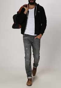 Zwarte ritssluiting hoodie, witte T-shirt, versleten grijze spijkerbroek en tan kleurige veterschoenen. Draagt een browngescheurde leren schoudertas.