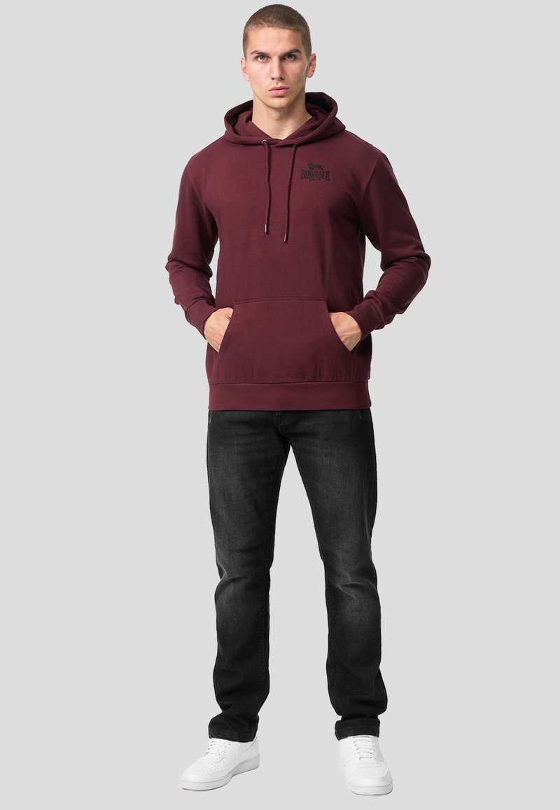 Homme debout portant un sweat à capuche bordeaux, jean noir, baskets blanches, mains dans les poches du sweat à capuche, fond gris neutre.