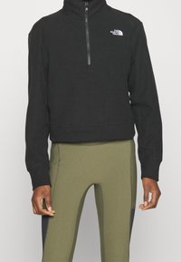 Schwarzer Fleece-Quarter-Zip-Pullover mit Stehkragen und einem weißen Logo auf der Brust, kombiniert mit olivgrünen, eng anliegenden Hosen.