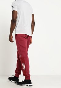 Maroon träningsbyxor med vitt tre-stripes mönster på sidan, parat med en grå T-shirt. Skorna är svarta träningsskor.