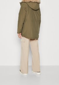 Olivově zelená parka s odnímatelnou kapucí s umělým kožíškem, zdůrazněným střihem v pase, bočními kapsami a volným střihem. Kombinováno s béžovými cargo kalhotami.