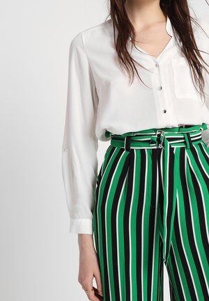 Vrouw draagt een witte blouse met knopen en een hoge taille broek met verticale strepen in groen, zwart en wit, met een bijpassende riem.