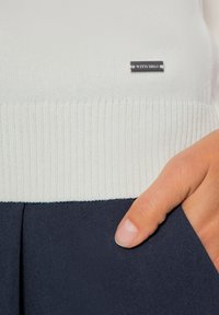 Hand met duim in een donkerblauwe zak onder een wit geribbelde breisweater met een klein metalen label waarop "Wittchen" staat.