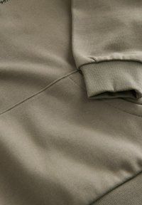 Olíva zöld pulóver anyag ribbed mandzsettákkal és sima felülettel. Vállain és testén varratok találhatók a strukturált megjelenés érdekében.