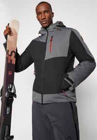 Chaqueta de esquí en negro y gris. Cuenta con un cierre frontal con cremallera, capucha ajustable, tejido texturizado y acentos rojos. Equipamiento de esquí sostenido en la mano izquierda.
