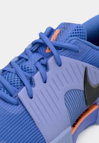 Μωβ αθλητικό παπούτσι με υφασμάτινο διχτυωτό επάνω μέρος, επίπεδα κορδόνια και φαρδιά ταινία σύσφιξης. Διαθέτει μαύρο λογότυπο και πορτοκαλί λεπτομέρεια.