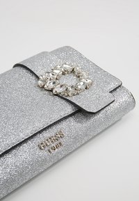 Portefeuille à paillettes argentées avec un rabat rectangulaire, orné d'un groupe ovale de pierres précieuses claires et de l'inscription "GUESS 1981" en lettres dorées.