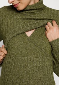 Femme soulevant la couche avant d'un pull côtelé vert olive, révélant un pull à col en V en dessous de la même couleur et tissu.
