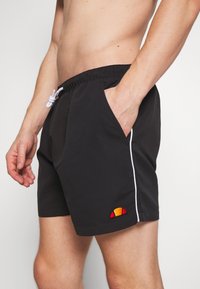 Bañador negro con cordón blanco, bolsillos laterales y un pequeño parche de logotipo multicolor cerca del dobladillo. Tejido suave y corte recto.