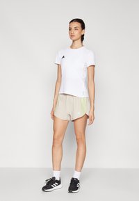 Camiseta deportiva blanca con logotipo negro en el pecho, combinada con pantalones cortos beige con rayas verdes y azules, llevada con zapatillas negras.