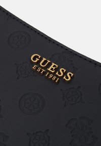 Černá kožená plocha s vyraženými kruhovými logy a zlatými kovovými písmeny "GUESS EST 1981" prošitými podél okraje.