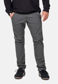 Mann in grauen Slim-Fit-Hosen und schwarzen Sneakers, der mit einer Hand in der Tasche vor einem einfarbigen Hintergrund steht.