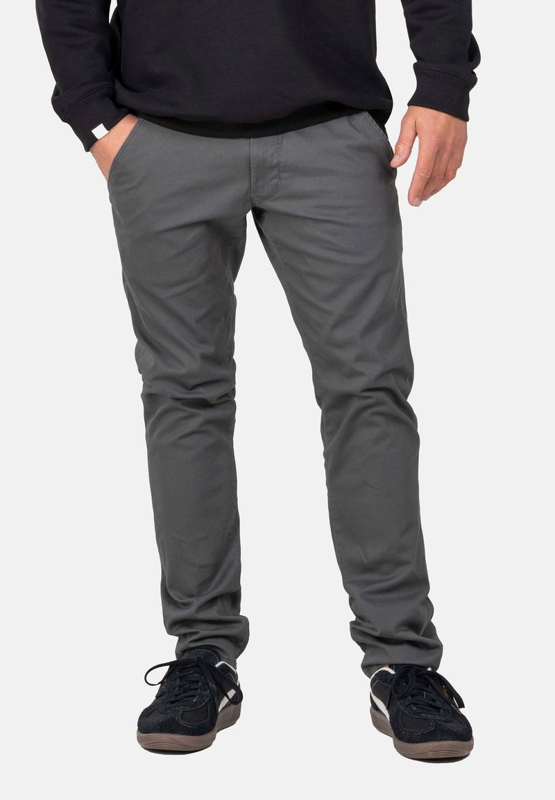 Mann in grauen Slim-Fit-Hosen und schwarzen Sneakers, der mit einer Hand in der Tasche vor einem einfarbigen Hintergrund steht.