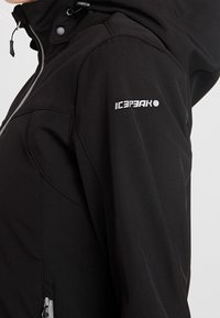 Veste softshell noire avec capuche, fermeture éclair visible et logo "Icepeak" sur le haut de la manche, portée par une personne montrant le haut du torse et le bras.