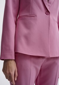 Růžový saka v hladké tkanině, s jedním knoflíkem a klopou, s elegantním detailem kapsy. Přiléhavý střih se strukturovanými rameny.