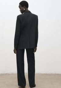Costume ajusté gris foncé avec une longue veste et un pantalon à jambes larges, présentant une texture lisse et des détails subtils de boutons.