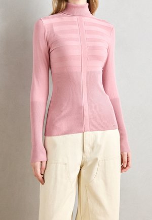 Personne portant un pull à col roulé rose ajusté en maille côtelée avec une texture rayée sur la poitrine et un pantalon large crème.