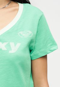 Πράσινο κοντομάνικο T-shirt με V-λαιμόκοψη, λευκό περίγραμμα και μερικό λευκό κείμενο, φορεμένο από άτομο με ορατό κολιέ αλυσίδα στον ώμο και τον λαιμό.