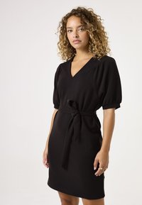 Vestido negro con escote en V, mangas abullonadas y cinturón atado en la cintura. Textura suave y silueta ajustada, diseño hasta la rodilla.